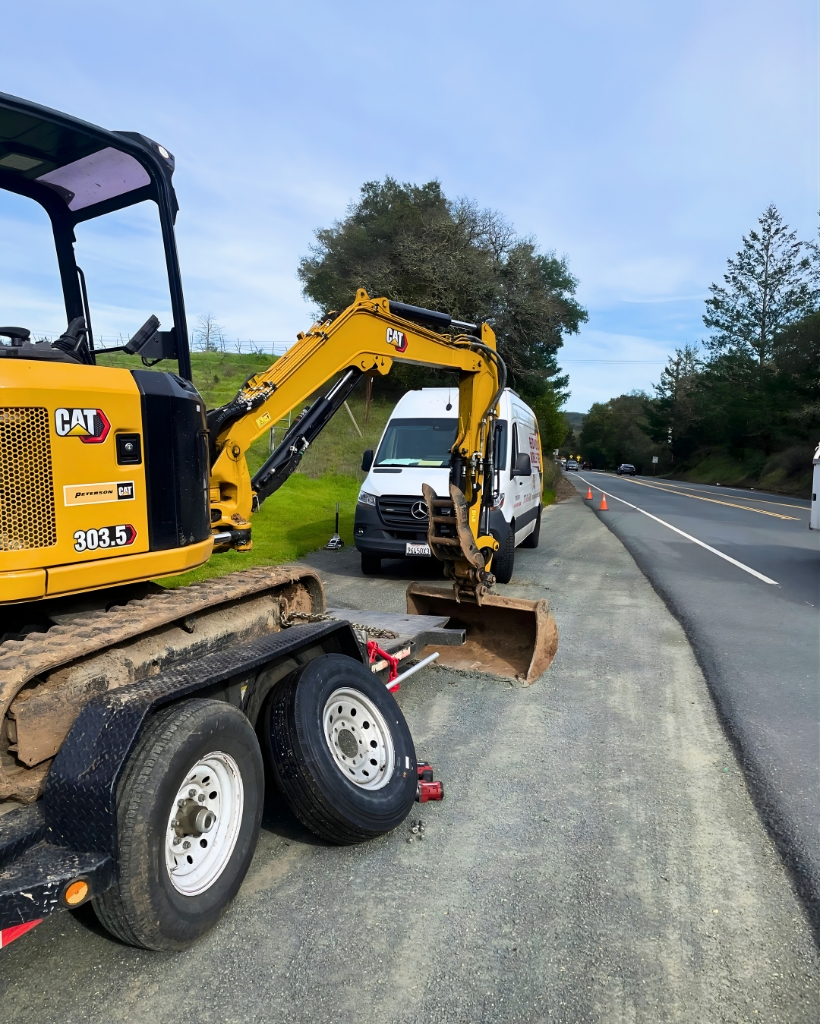 Yellow cat excavator mounted on a trailer parked on a gravel roadside beside a two-lane road, with a white van in the background and orange cones ahead.