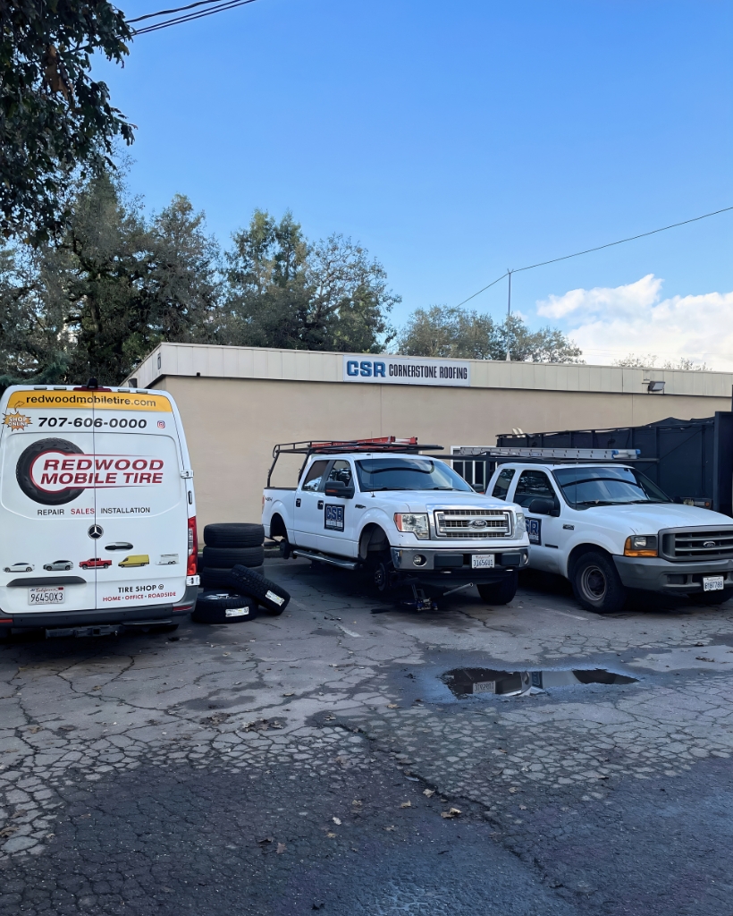 A small auto shop lot with three white service vehicles, tires piled on the ground, and a sign for csr cornerstone roofing on the building behind them.