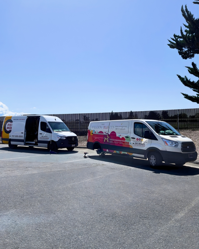 Two white service vans with colorful graphics parked in a sunny lot; one van's side door is open.