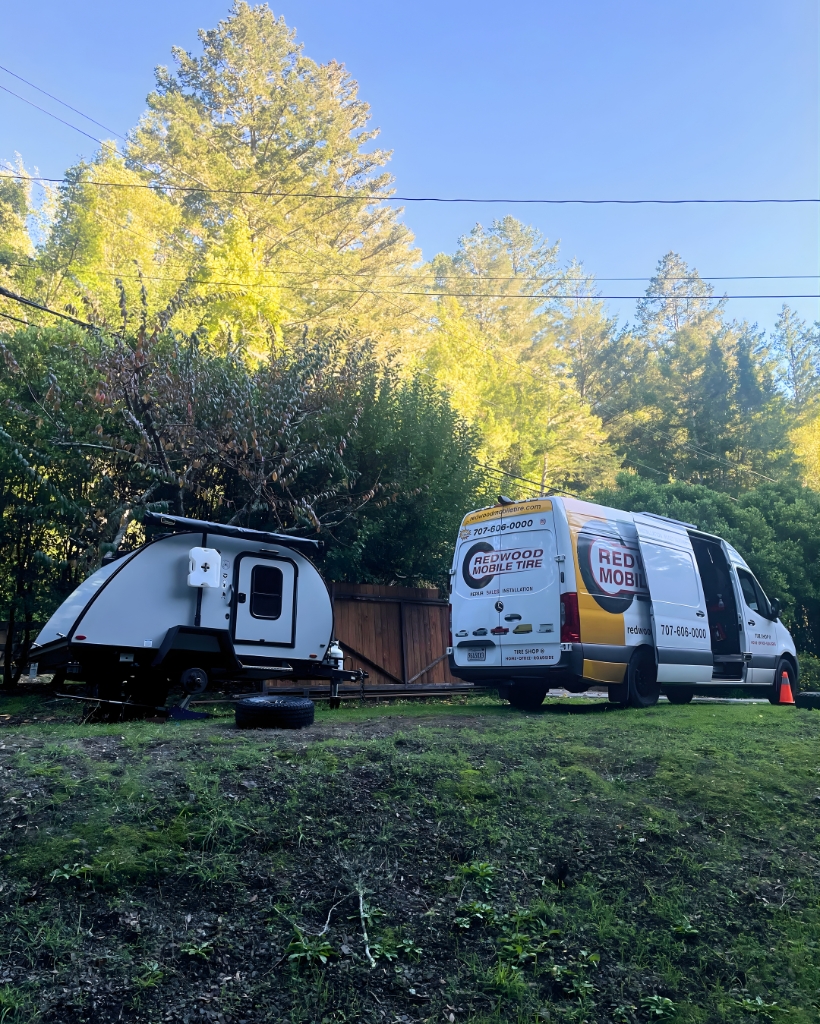 White teardrop camper hitched to a small trailer beside a wooden fence in a grassy yard, with a branded service van (redwood mobile tire) nearby.