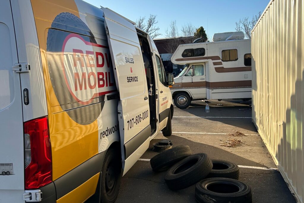White service van with its side door open reading 'sales & service', parked beside a stack of used tires in a lot with an rv in the background.